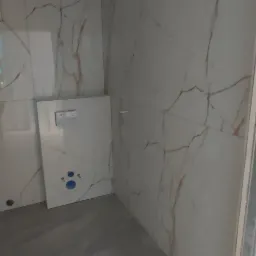 Wnętrze łazienki w trakcie wykańczania, widoczne płytki imitujące marmur na ścianach, szara posadzka i zamontowany stelaż podtynkowy WC z widocznymi przyłączami.