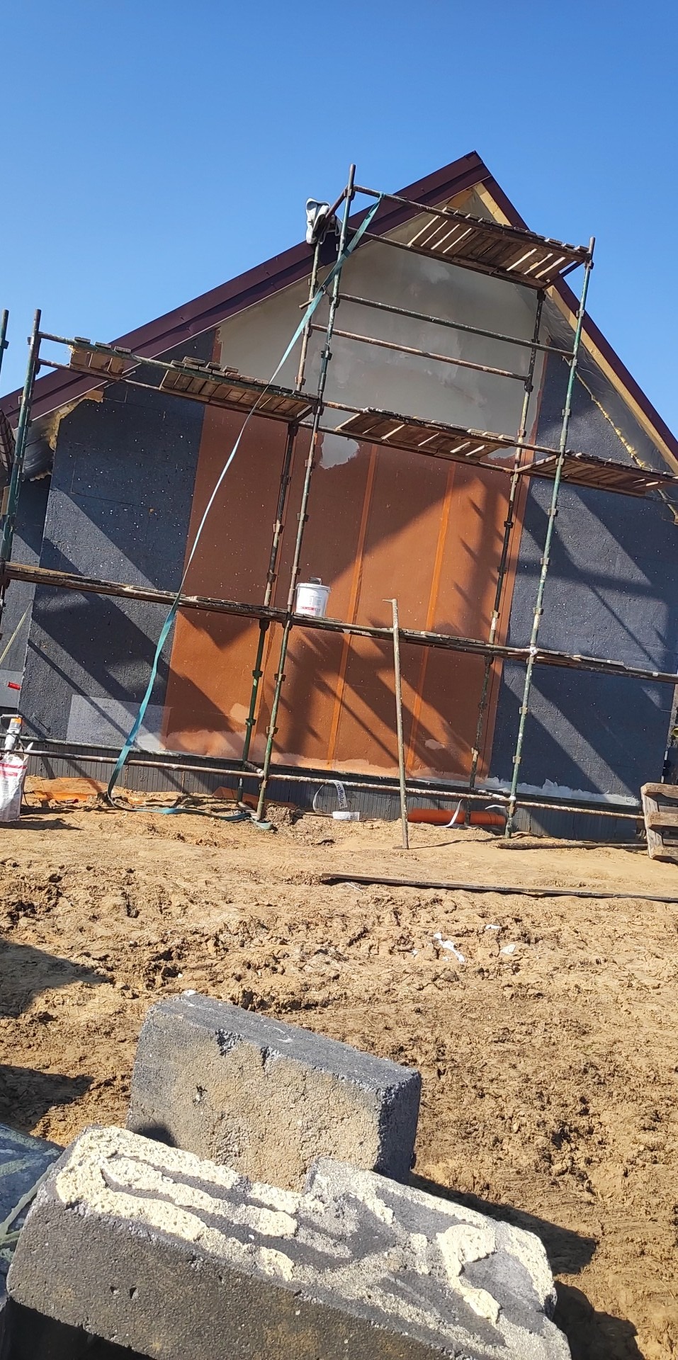 Dom w trakcie ocieplania, widoczne rusztowania, warstwa izolacyjna i częściowo nałożony tynk w kolorze brązowym, na pierwszym planie kostka brukowa.