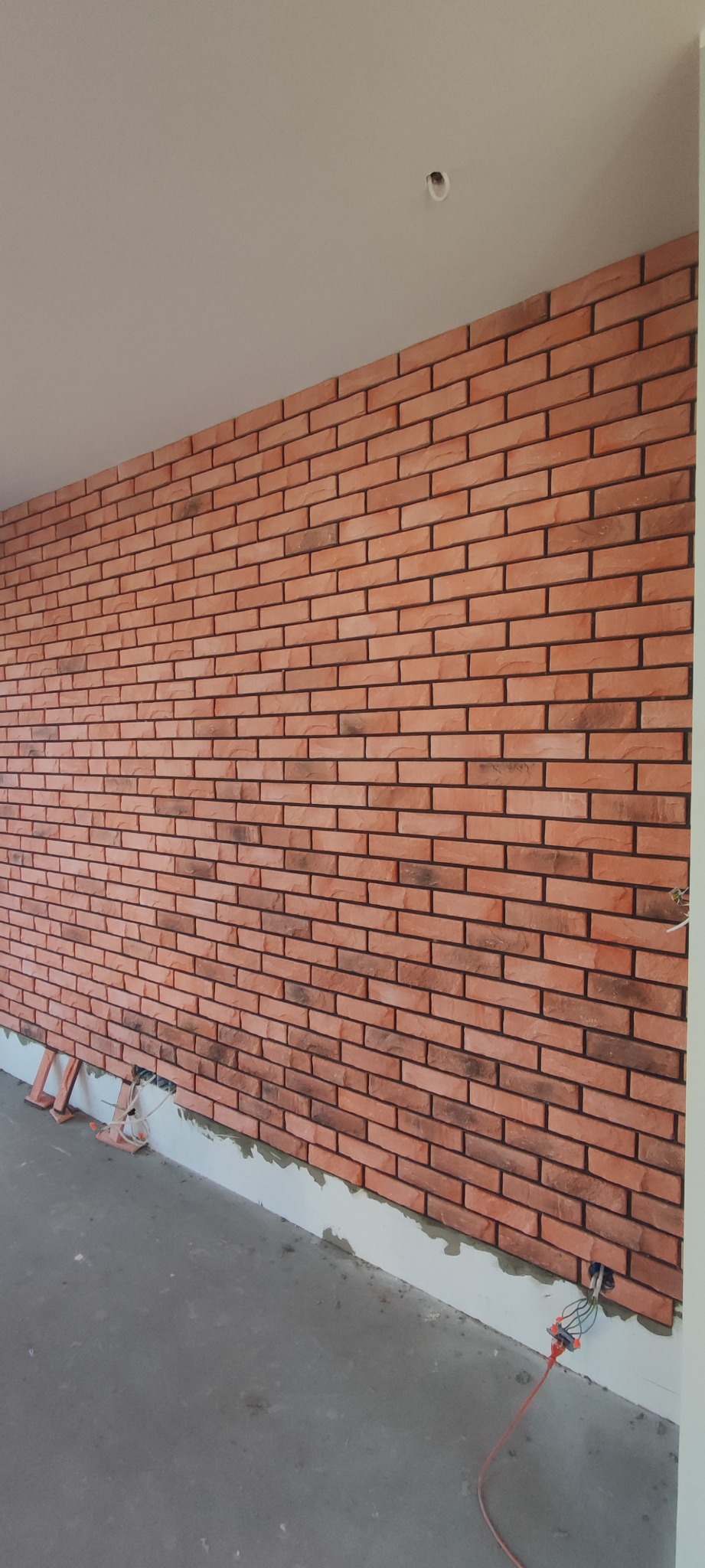 Ściana wyłożona dekoracyjną cegłą w odcieniach czerwieni i brązu, widoczne przewody elektryczne przy podłodze, mała drabinka oparta o ścianę, szara, surowa podłoga, biały sufit.