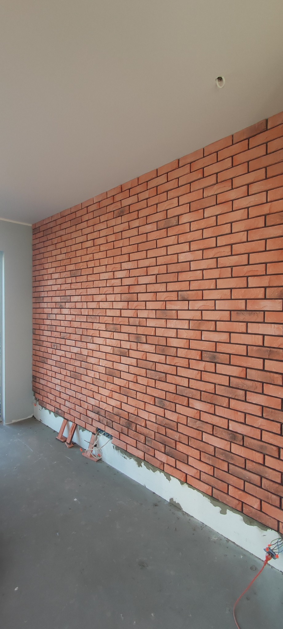 Ściana wyłożona cegiełkami w odcieniach czerwieni i brązu, widoczne przewody elektryczne wystające ze ściany, podłoga w stanie surowym, fragment okna po lewej stronie.