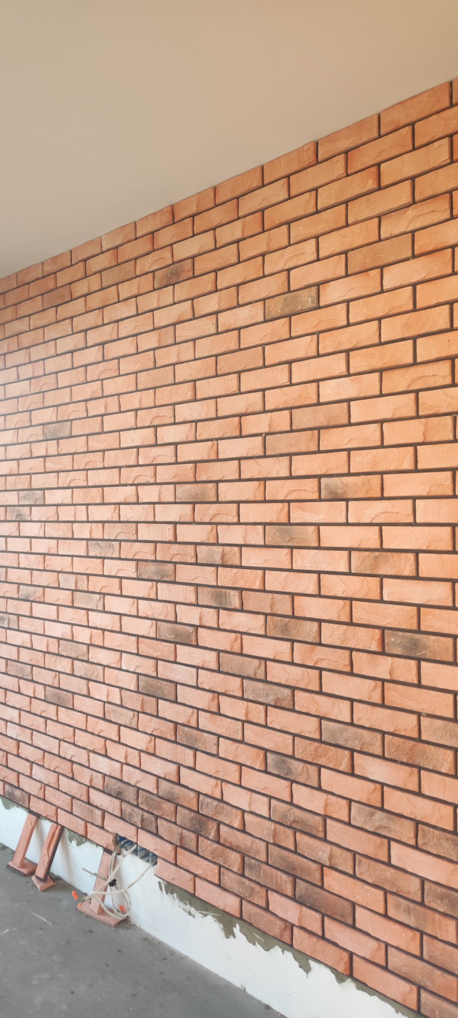 Ściana wykończona cegiełkami klinkierowymi w odcieniach pomarańczy i brązu, widoczne fragmenty instalacji elektrycznej i podpórki montażowe przy podłodze.