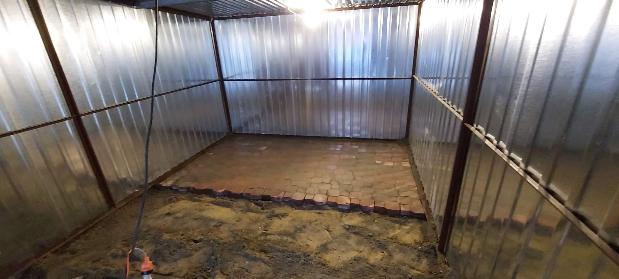 Wnętrze metalowego garażu z częściowo ułożoną kostką brukową na podłożu z piasku i ziemi, widoczne metalowe ściany i konstrukcja dachu.