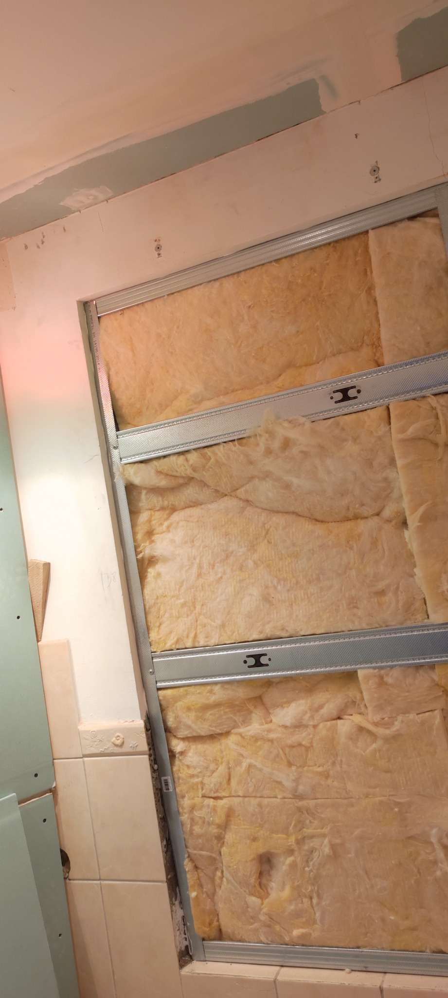 Izolacja z wełny mineralnej w metalowym stelażu, widoczne płytki ceramiczne i fragment płyty gipsowo-kartonowej podczas prac wykończeniowych w łazience.