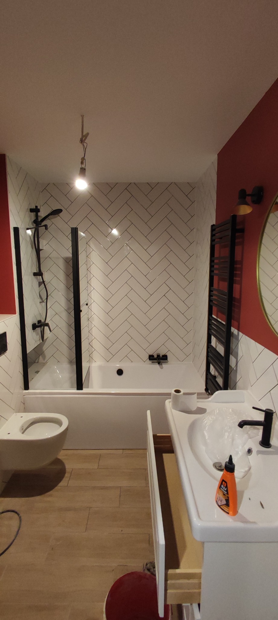 Nowoczesna łazienka z białą armaturą, kabiną prysznicową z czarnymi ramami, grzejnikiem drabinkowym i ścianą w biało-czerwone pasy, wykończona płytkami ułożonymi w jodełkę.