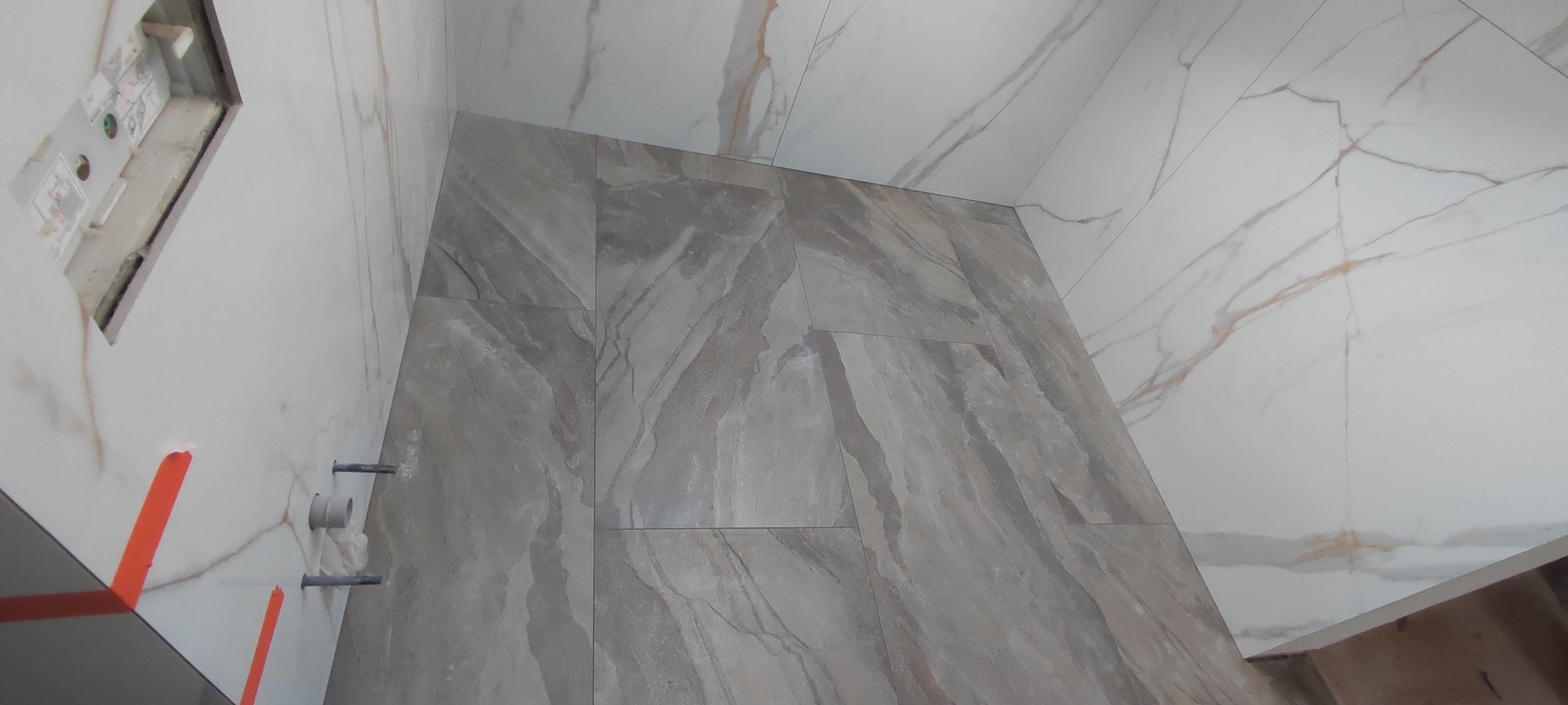 Fragment łazienki w trakcie wykańczania, widoczne płytki imitujące marmur na ścianach, zamaskowane rury oraz otwór na instalację elektryczną.