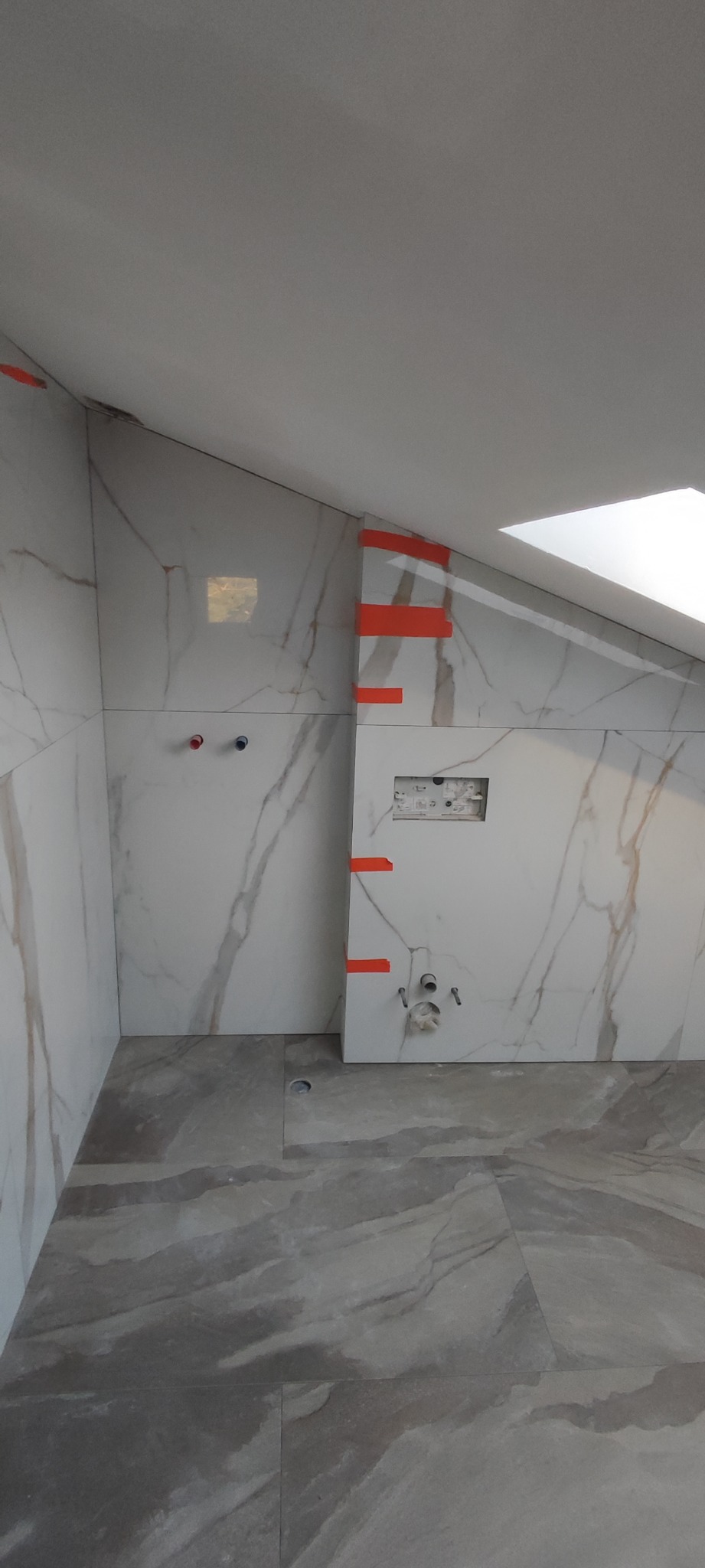 Łazienka w trakcie wykańczania: białe płytki imitujące marmur na ścianach, szare płytki podłogowe, widoczne otwory na rury i instalacje, zabezpieczone pomarańczową taśmą. Skos dachu z oknem...