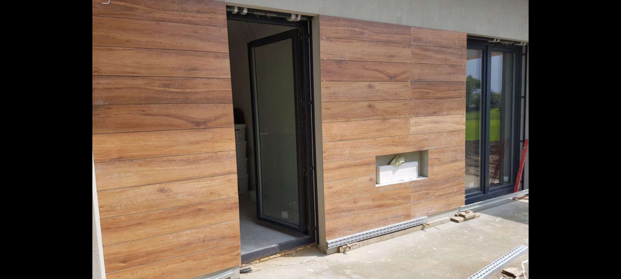 Wykończenie elewacji budynku panelami drewnopodobnymi z otwartymi drzwiami balkonowymi i oknem, widoczne elementy montażowe i fragment elewacji z pustką wypełnioną styropianem.