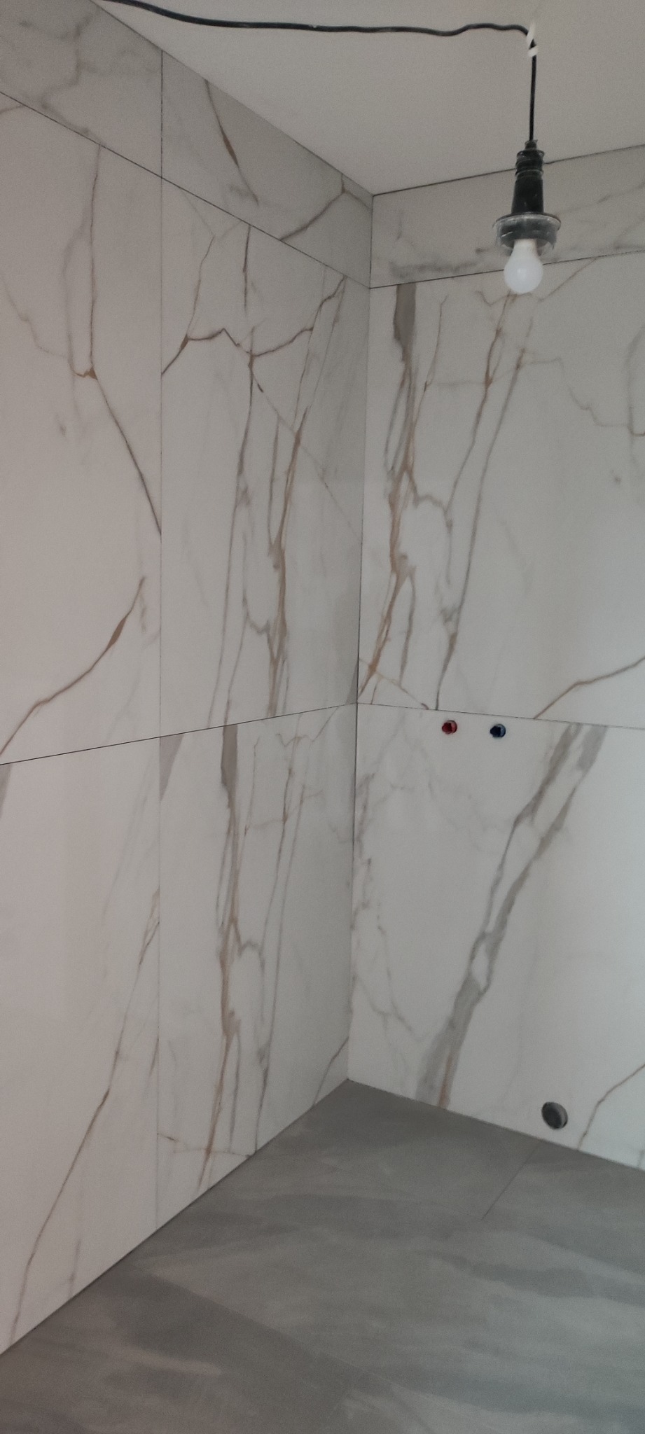 Wykończona ściana w łazience z białych płytek imitujących marmur, szara podłoga, widoczne przyłącza wodne i prowizoryczna lampa wisząca.