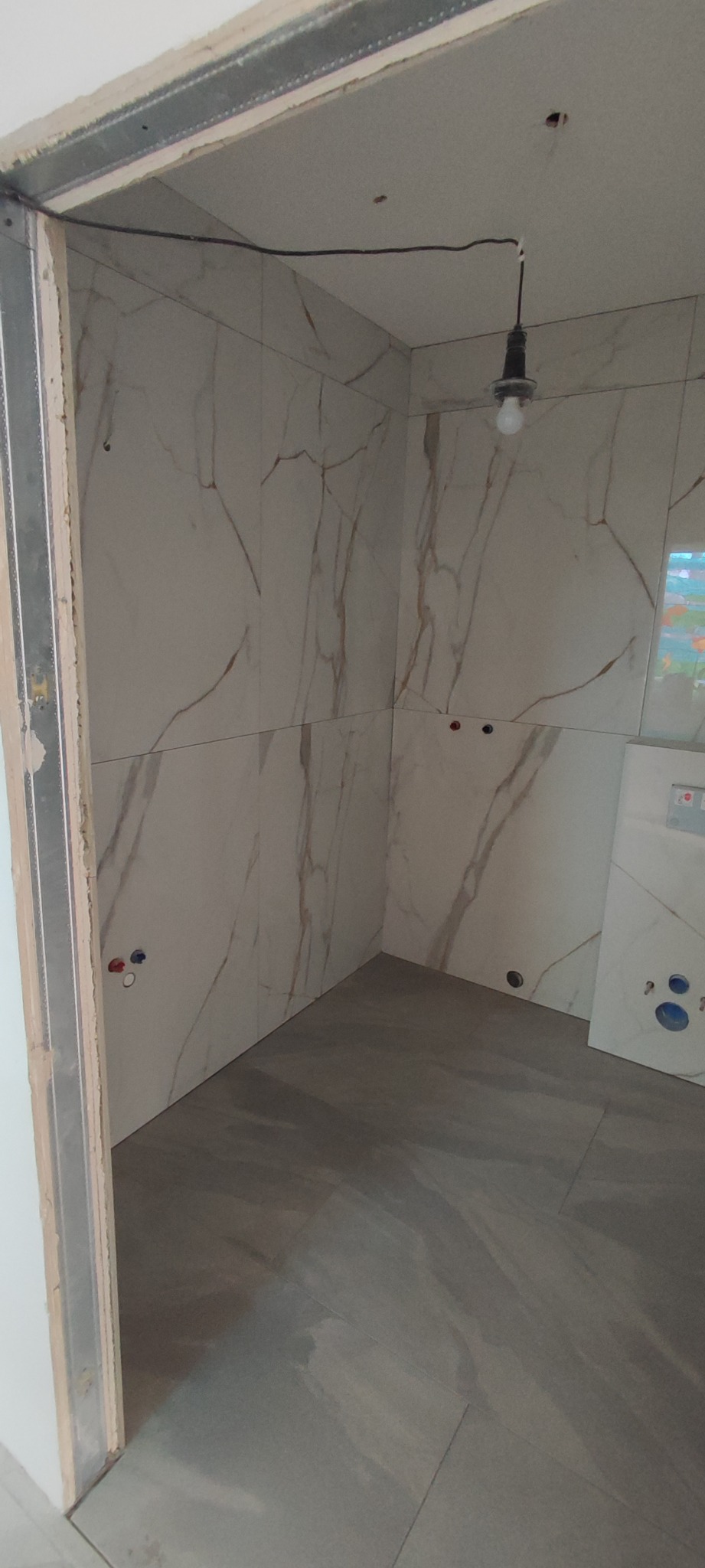 Wnętrze łazienki w trakcie wykończenia: marmurowe płytki ścienne z widocznymi przyłączami wodno-kanalizacyjnymi, surowa żarówka na kablu, podłoga w szarych płytkach imitujących kamień, metalowa...