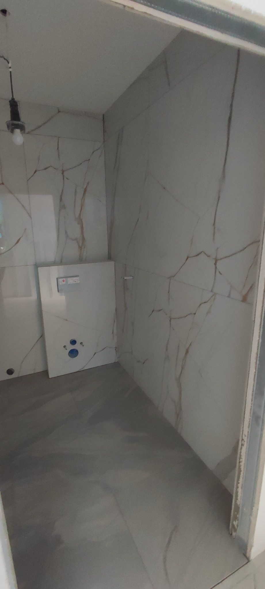 Wnętrze łazienki w trakcie wykańczania, widoczne płytki imitujące marmur na ścianach, szara wylewka na podłodze i zamontowany stelaż podtynkowy WC z widocznymi otworami na przyłącza.