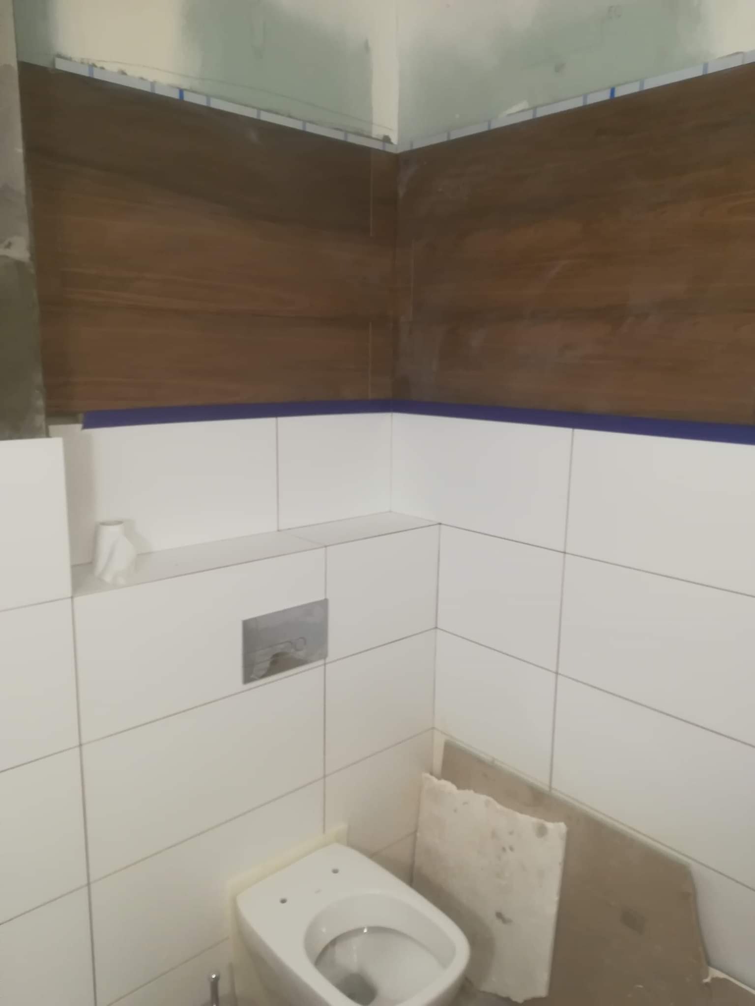 Wnętrze łazienki w trakcie wykańczania, widoczne białe płytki ceramiczne na ścianach, częściowo wykończone brązowe panele imitujące drewno, wisząca muszla klozetowa i fragmenty materiałów...