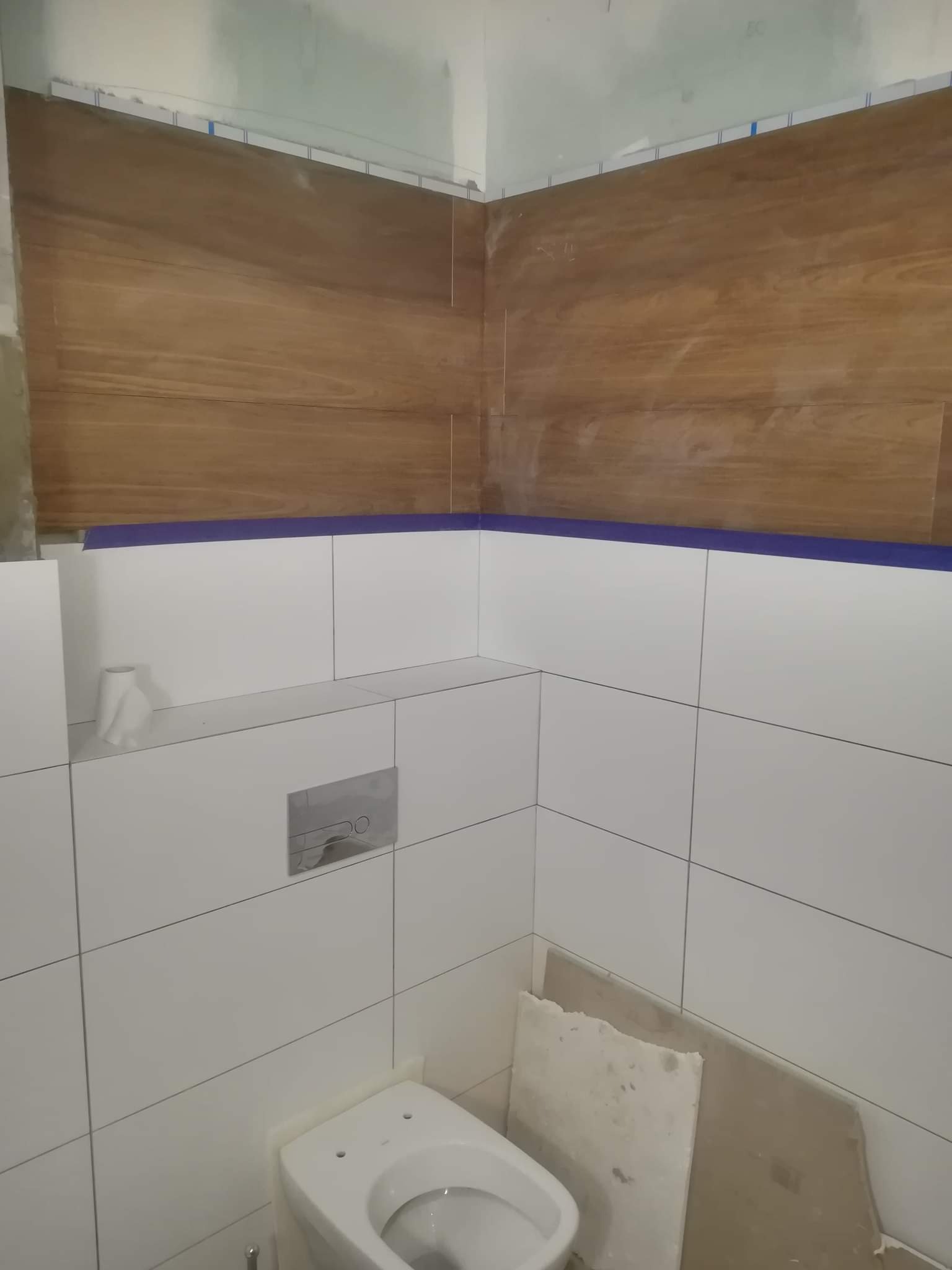 Wnętrze łazienki w trakcie prac wykończeniowych, widoczne białe płytki na ścianach, drewnopodobne płytki u góry, niebieska taśma malarska, częściowo zamontowana toaleta i płyty gipsowe na podłodze.