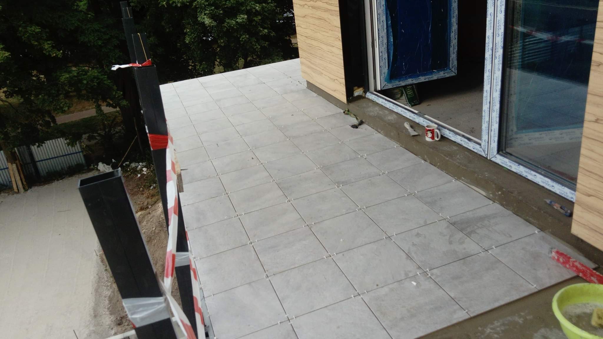 Układanie szarych płytek na tarasie, widoczne krzyżyki dystansowe, okno tarasowe z folią ochronną, kubełek z wodą i gąbką, fragment ogrodzenia w tle.