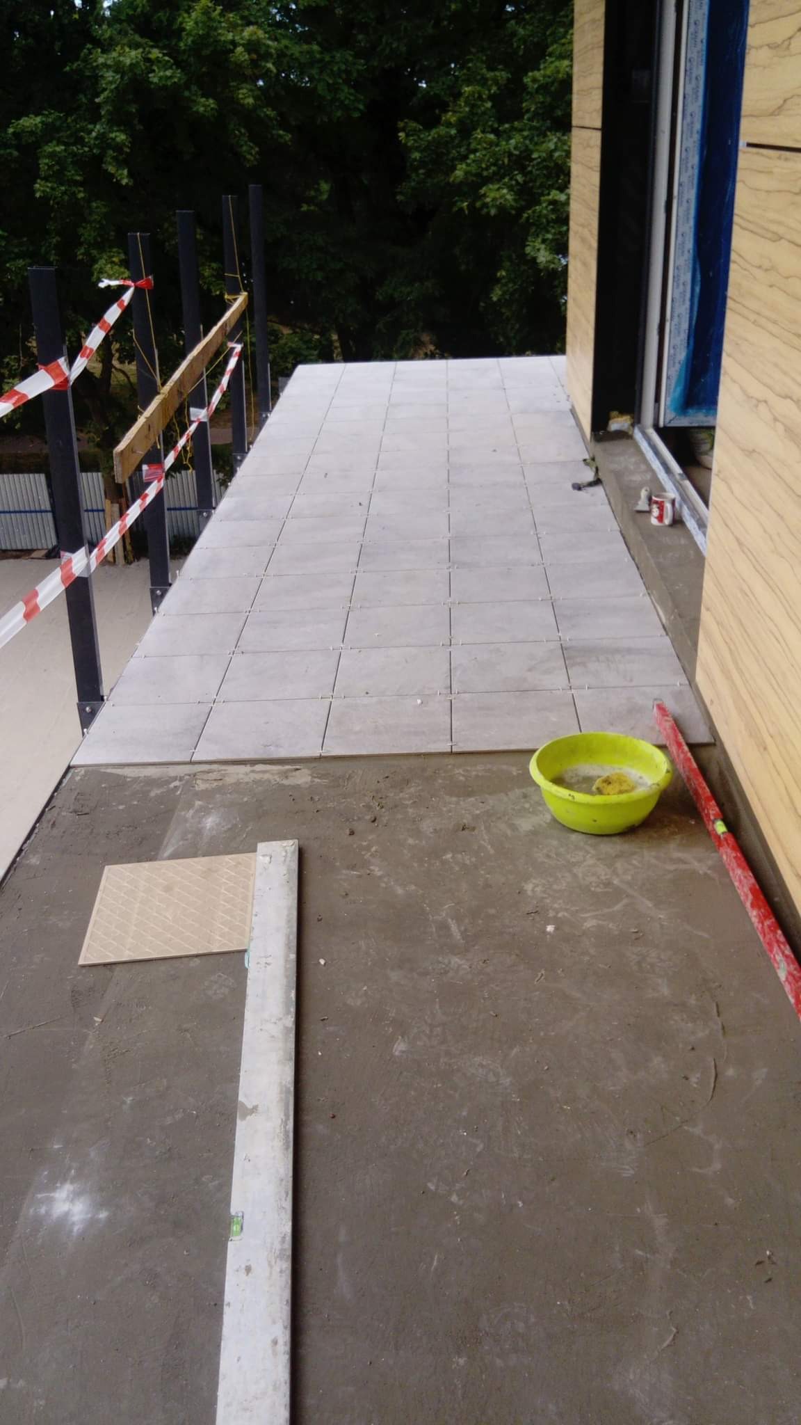 Montaż jasnoszarych płytek na tarasie z widocznym poziomem, miską z wodą i fragmentem niezakończonej powierzchni oraz barierką z taśmą ostrzegawczą.