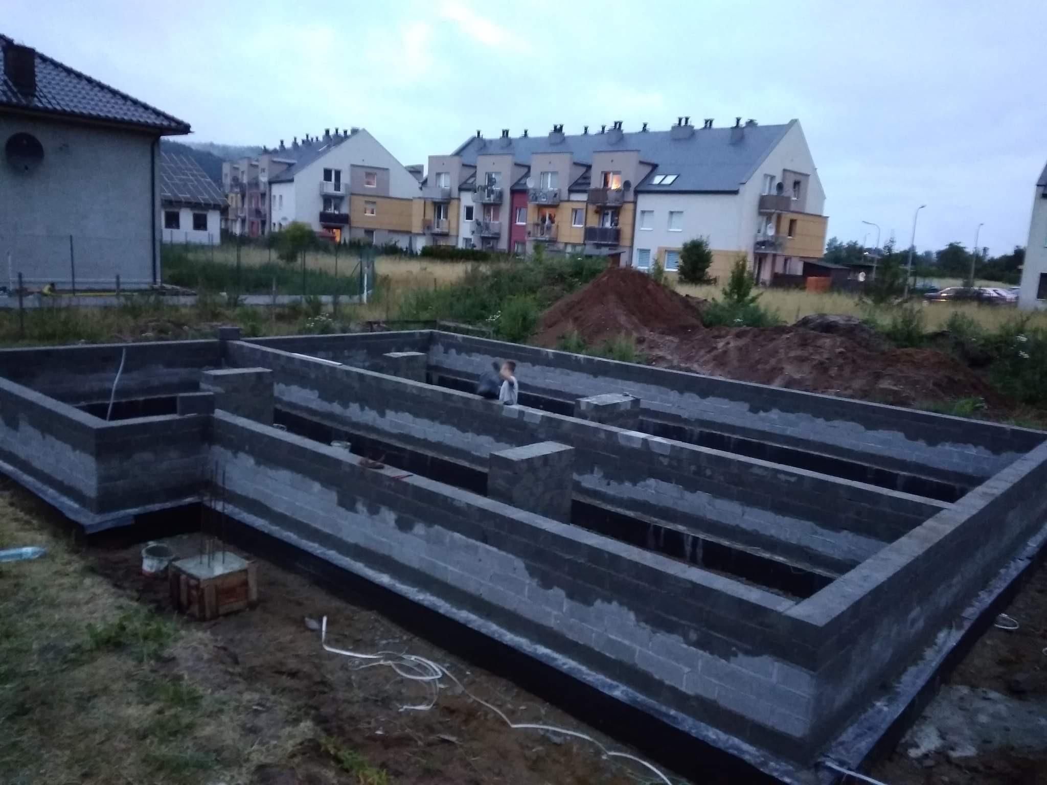 Wykop pod fundamenty domu jednorodzinnego, częściowo wylane ściany z szarego bloczka betonowego, widoczne zbrojenia, w tle zabudowa wielorodzinna i hałda ziemi.