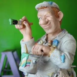 Figurka uśmiechniętego malarza z pędzlem i wiadrem farby na tle zielonej ściany, z drabinką w tle.