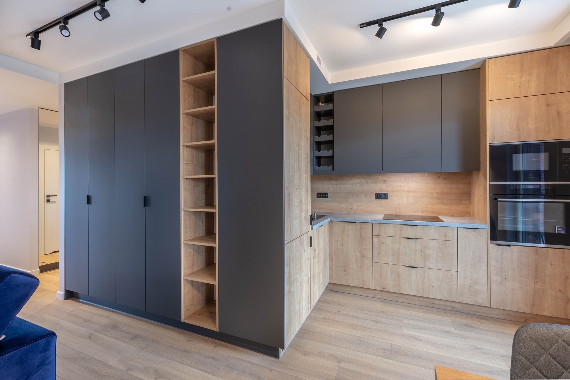 Nowoczesna kuchnia z zabudową meblową: połączenie forniru dębowego i grafitowych frontów, oświetlenie punktowe sufitowe, minimalistyczne uchwyty, zintegrowany regał z półkami, płyta indukcyjna...
