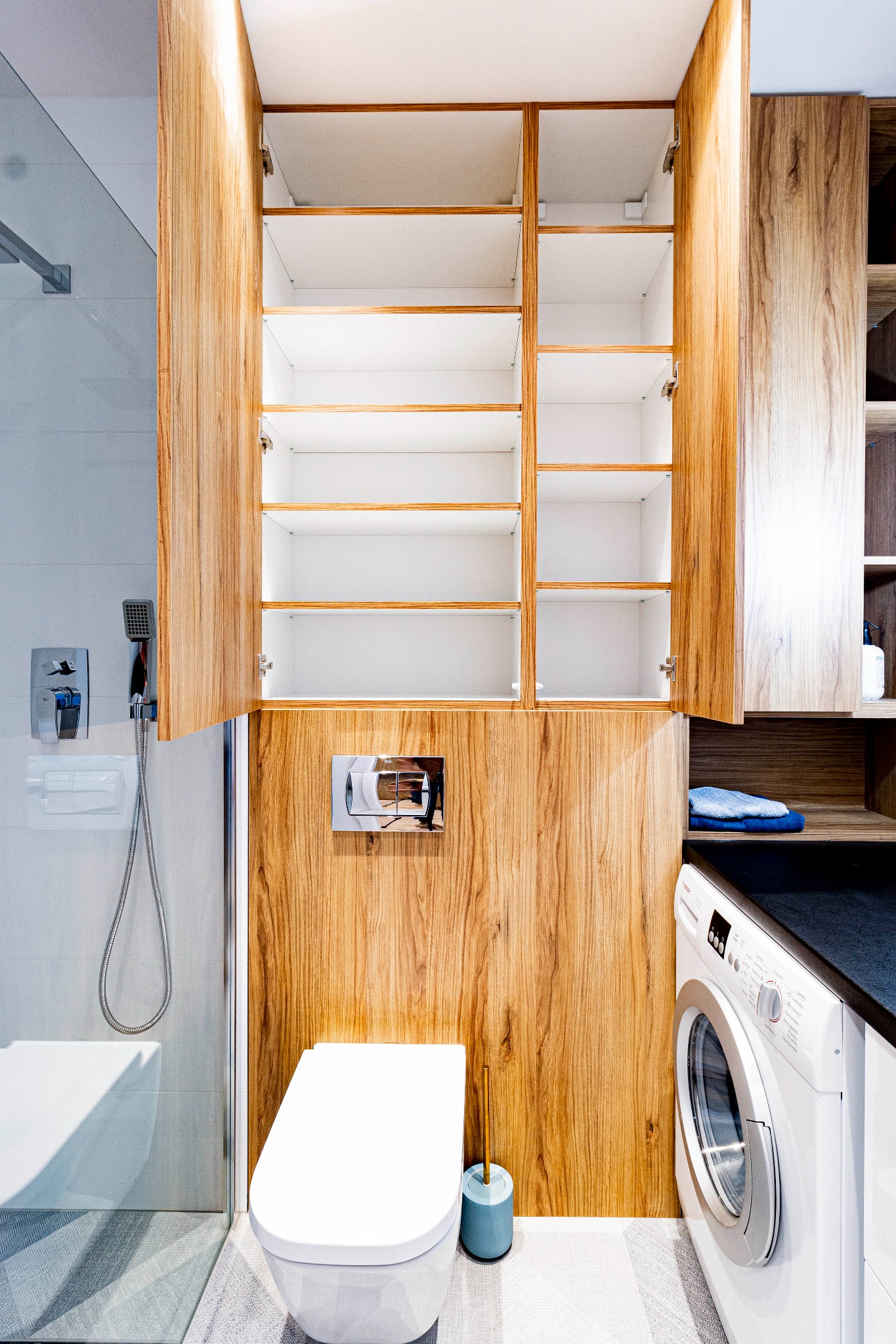 Nowoczesna łazienka z drewnianą zabudową, otwartymi szafkami nad toaletą, prysznicem ze szklaną kabiną i pralką Bosch z czarnym blatem.