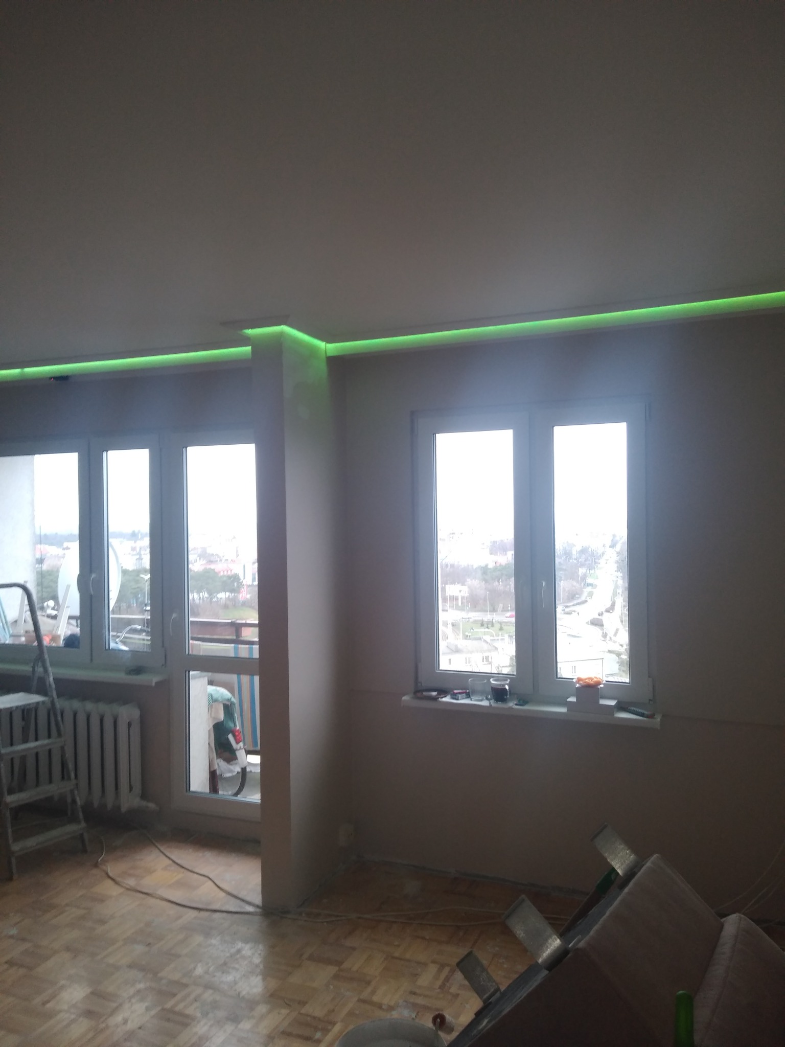 Pokój po remoncie z podświetlonym na zielono sufitem, widoczny fragment parkietu, drabina i okno z widokiem na miasto.