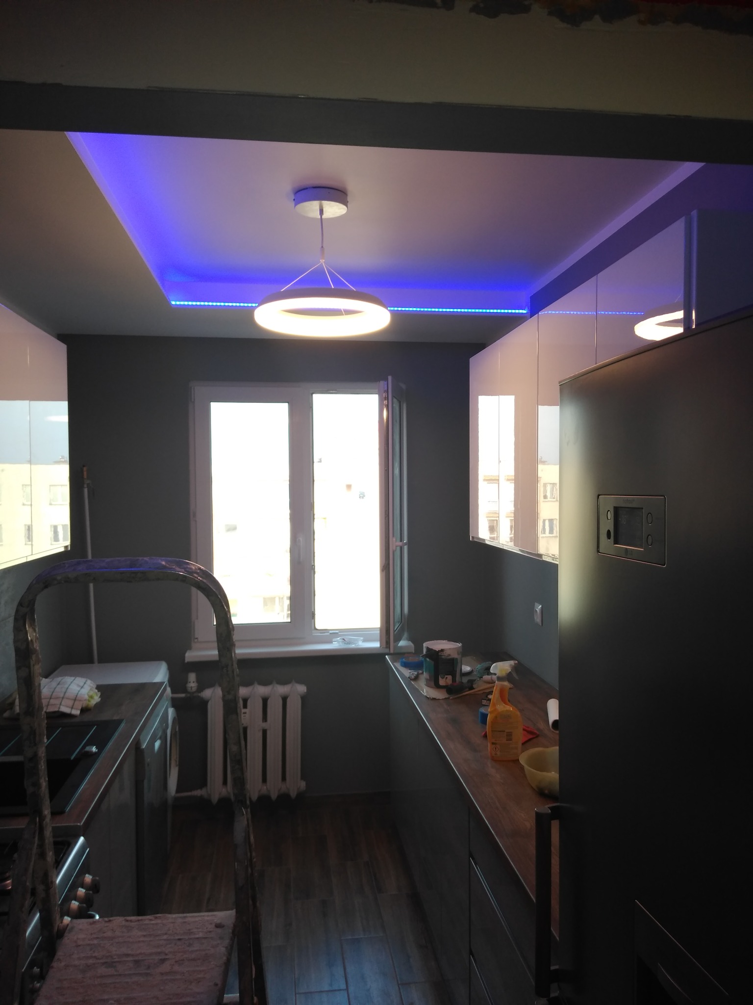 Wnętrze kuchni w trakcie remontu, widoczna drabina, okno, zabudowa kuchenna w szaro-białej kolorystyce, sufit podwieszany z oświetleniem LED w kolorze niebieskim oraz okrągła lampa wisząca.