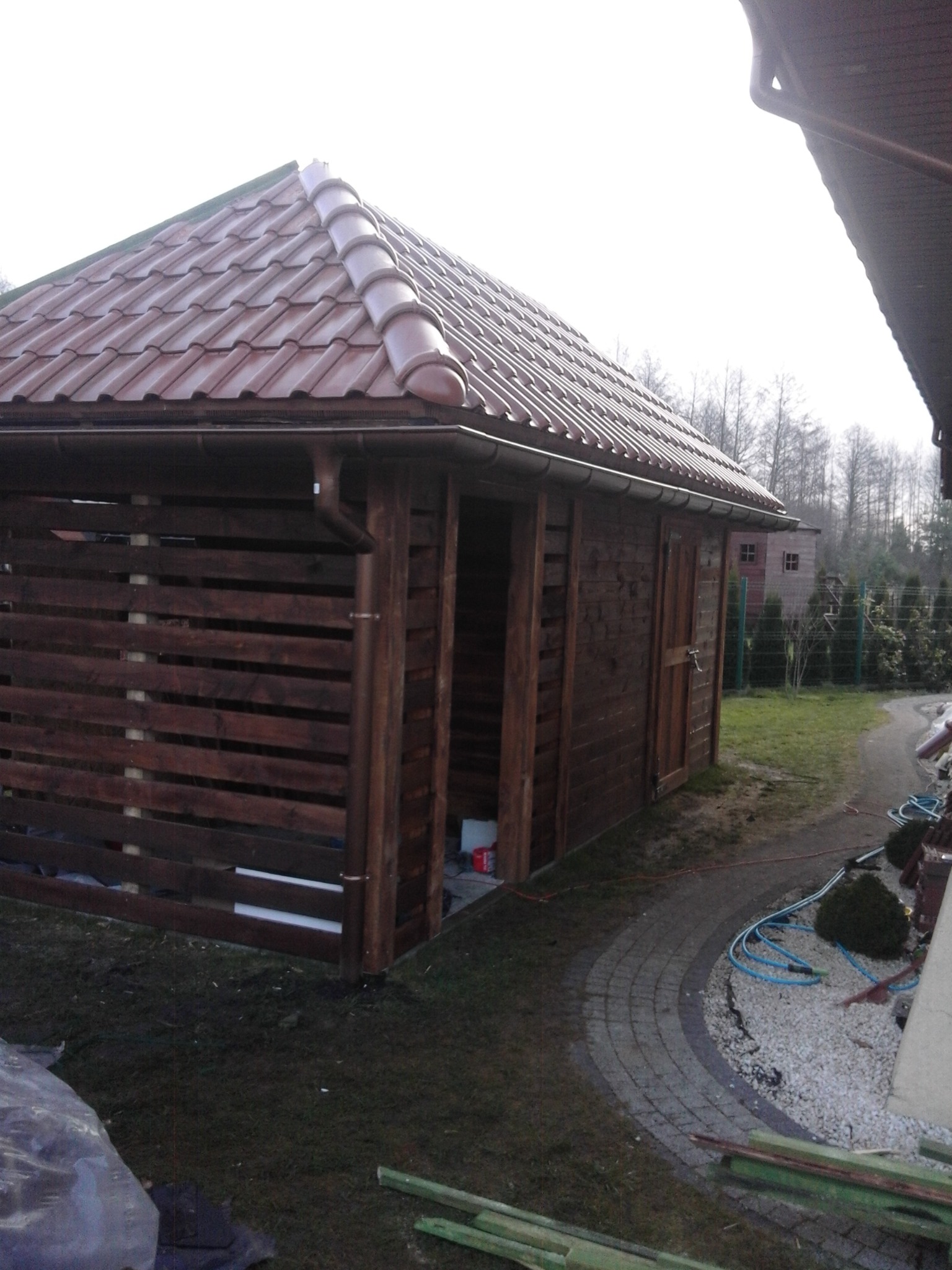 Drewniana wiata z brązową dachówką ceramiczną, widoczne orynnowanie, częściowo ażurowe ściany z desek, drzwi zamykane na zasuwkę, obok ścieżka z kostki brukowej i żwiru.