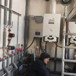 Instalacja dwóch kotłów grzewczych Ariston z podłączeniami hydraulicznymi i elektrycznymi, widoczni dwaj instalatorzy w trakcie pracy, rury grzewcze z izolacją, przewody elektryczne i drabina.