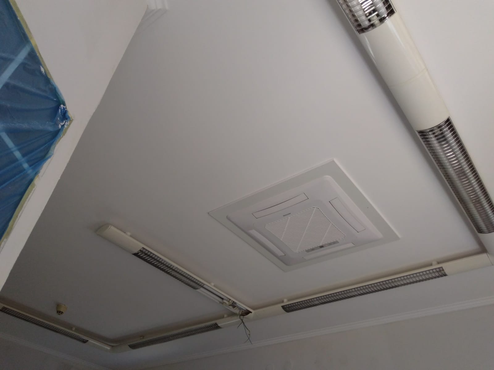Kasetonowa jednostka klimatyzacji zamontowana w suficie podwieszanym, widoczne również oprawy oświetleniowe i fragment zabezpieczonej folią ściany.