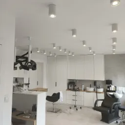 Jasne wnętrze salonu fryzjerskiego z licznymi punktowymi lampami sufitowymi, stanowiskami pracy z fotelami i umywalkami, minimalistycznymi białymi szafkami w tle oraz suszarką fryzjerską...