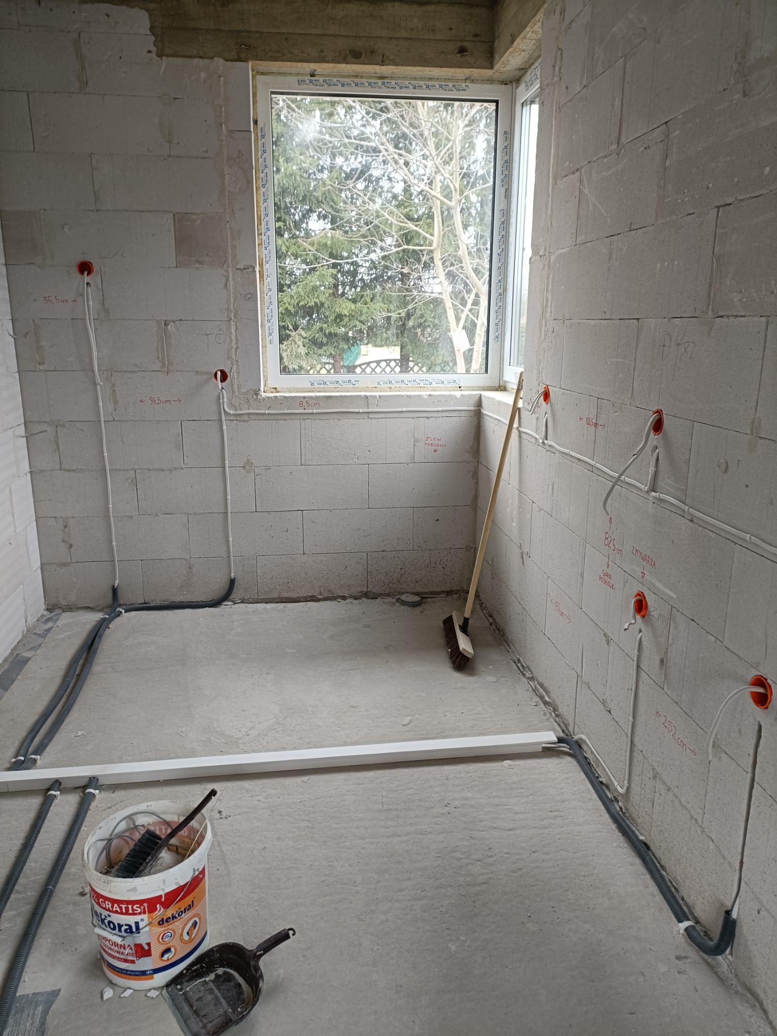 Wnętrze w trakcie prac wykończeniowych: surowe ściany z betonu komórkowego z widocznymi punktami elektrycznymi, peszle i przewody ułożone wzdłuż ścian, okno z widokiem na drzewa, wiadro...