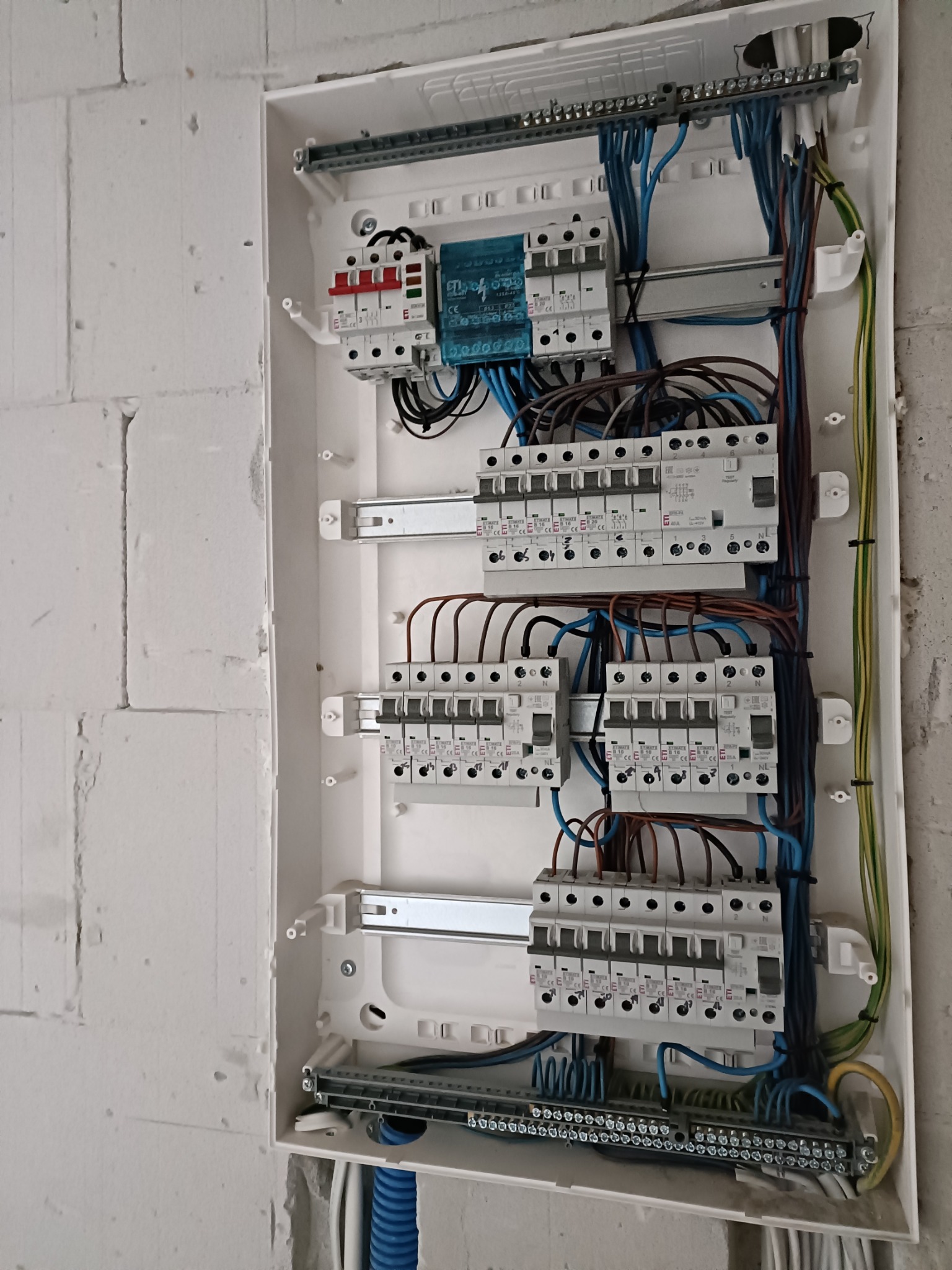 Rozdzielnica elektryczna z zamontowanymi bezpiecznikami i przewodami w surowym wnętrzu, widoczne ślady tynku na ścianie.