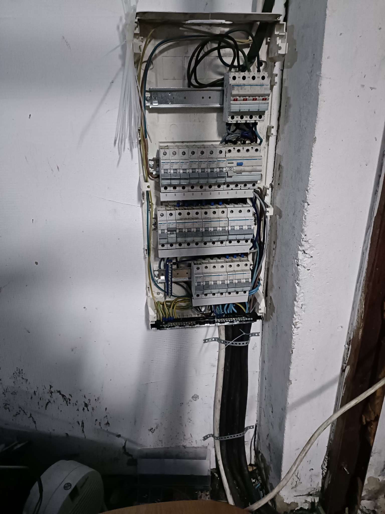 Otwarta skrzynka elektryczna z widocznymi bezpiecznikami i przewodami na tle ściany z odpryskującą farbą, zamocowana na metalowych szynach.