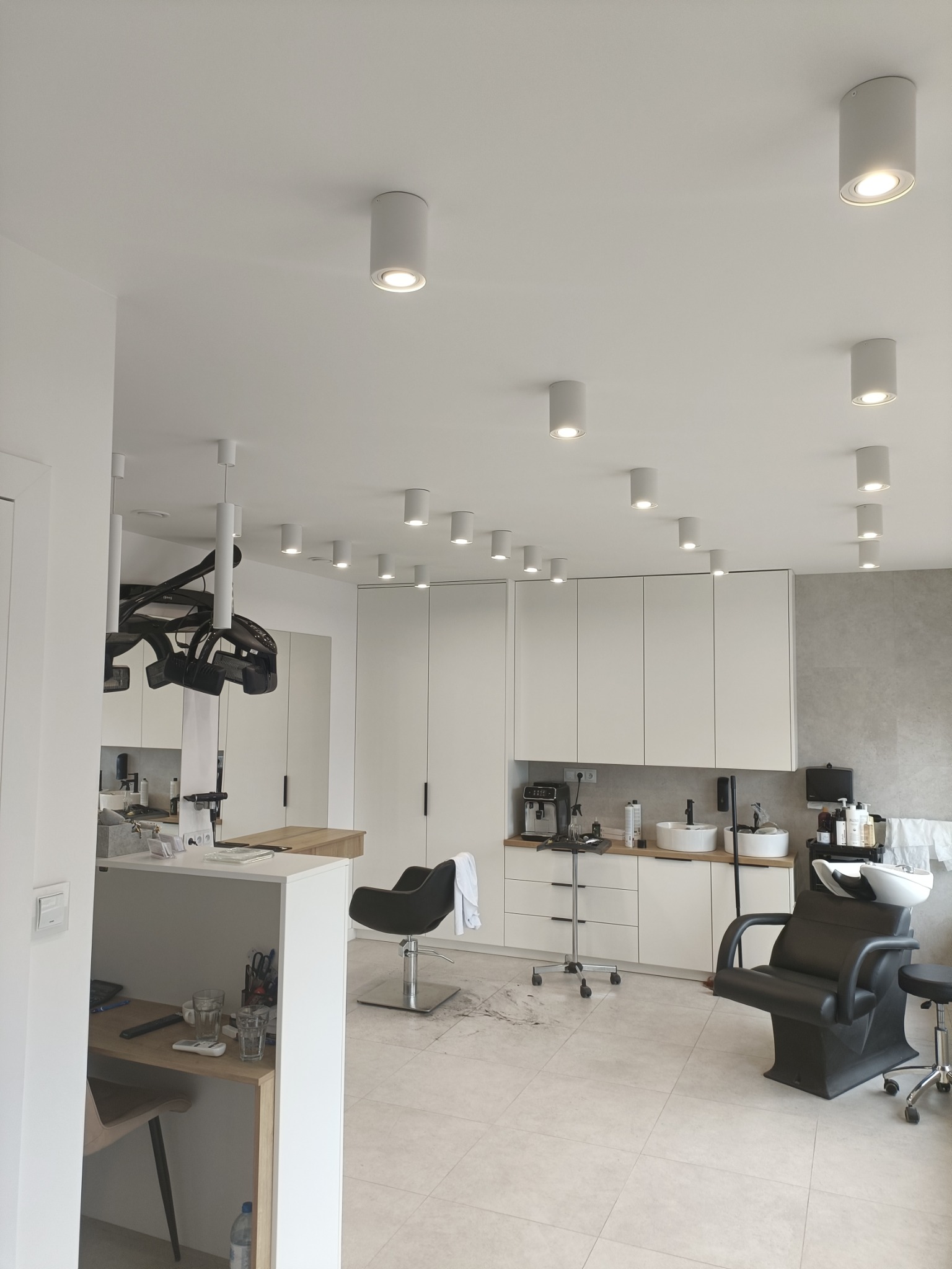 Jasne wnętrze salonu fryzjerskiego z licznymi punktowymi lampami sufitowymi, stanowiskami pracy z fotelami i umywalkami, minimalistycznymi białymi szafkami w tle oraz suszarką fryzjerską...