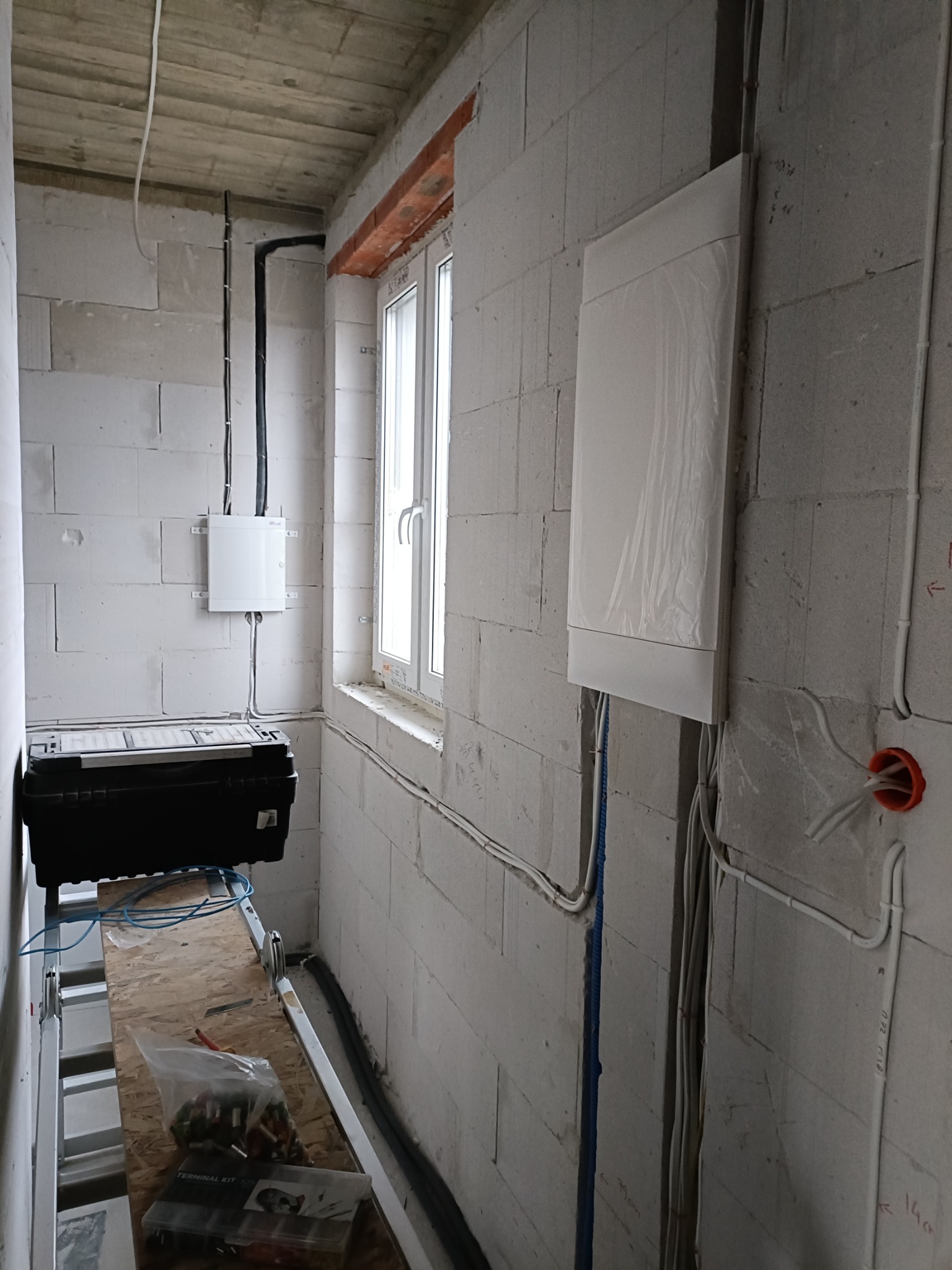 Wnętrze w trakcie prac elektrycznych: widoczne kable, puszki instalacyjne, skrzynka narzędziowa na drabinie, ściany z betonu komórkowego, okno.