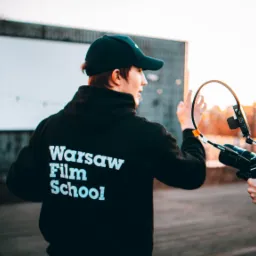 Ekipa filmowa podczas kręcenia ujęcia na zewnątrz, jeden z operatorów trzyma kamerę ze stabilizatorem, drugi w bluzie z napisem Warsaw Film School, w tle budynek z pustą tablicą.