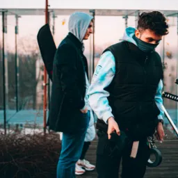 Kobieta trzymająca dwie katany na planie filmowym, obok mężczyzna z akcesoriami, w tle szklana konstrukcja i budynki.