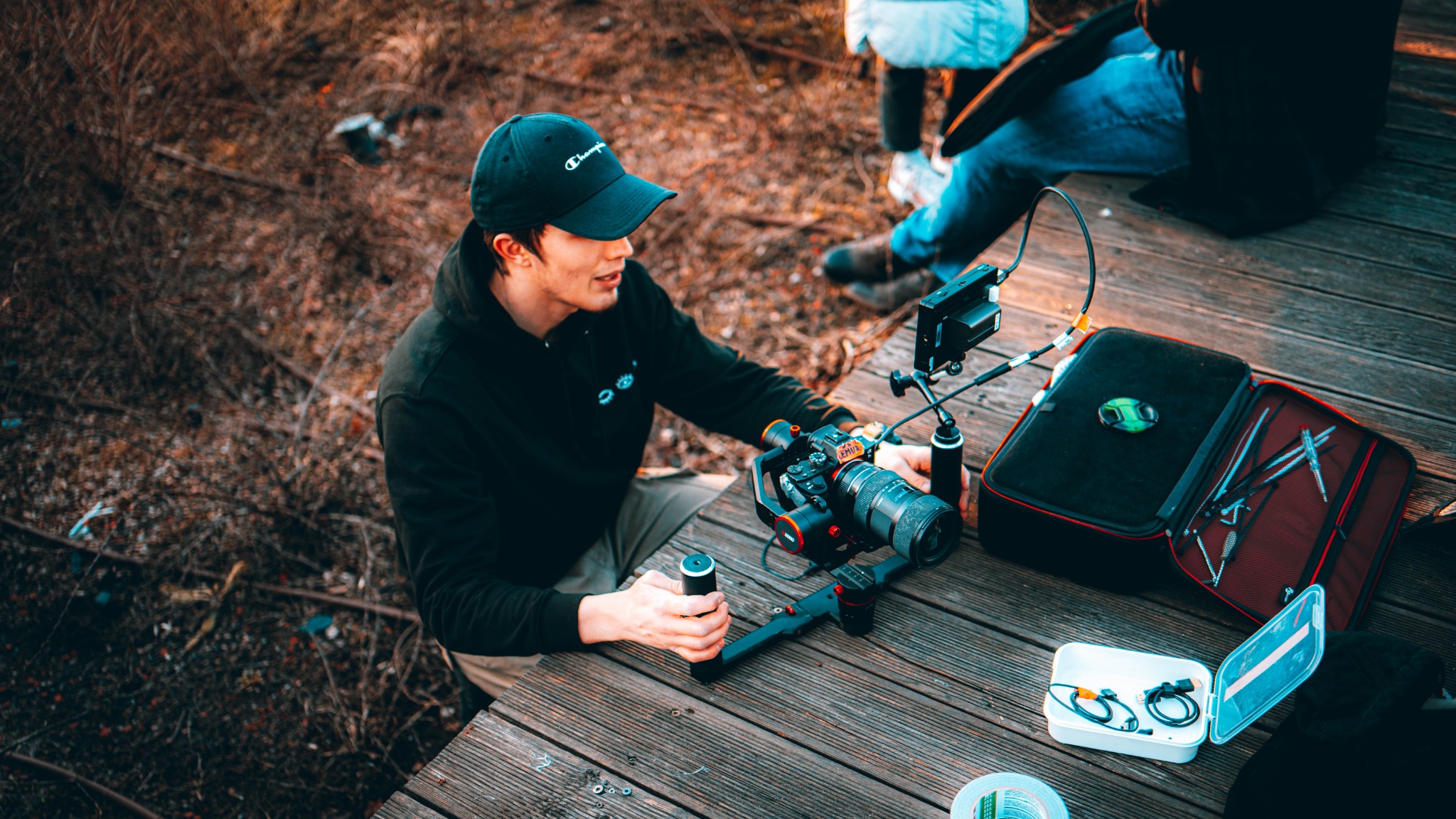 Młody mężczyzna w czarnej bluzie i czapce z daszkiem obsługuje profesjonalny stabilizator kamery z zamontowanym aparatem na drewnianym pomoście, obok leżą akcesoria w futerale i pudełku.