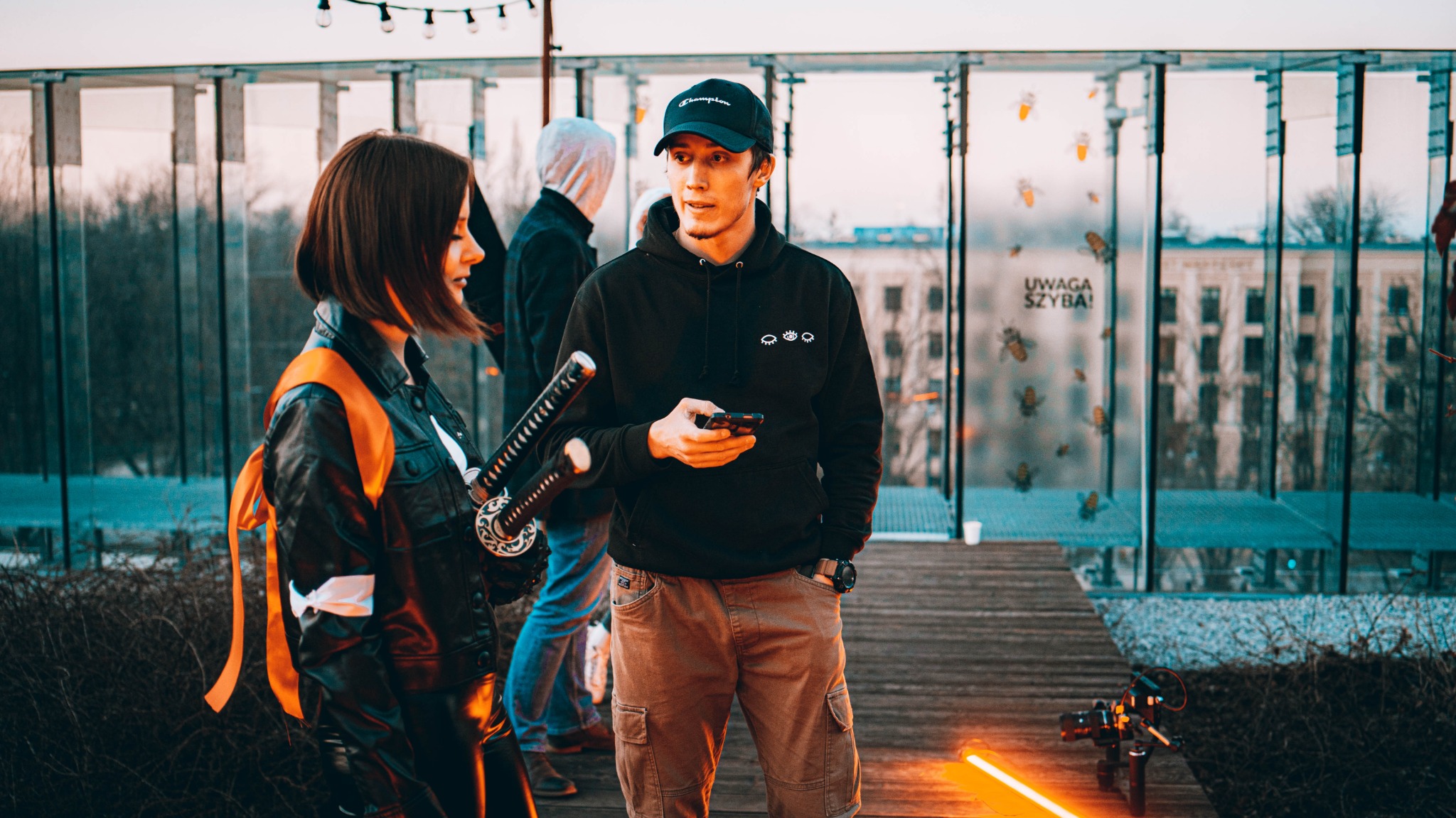 Ekipa filmowa w plenerze: kobieta w kostiumie z kataną, mężczyzna w bluzie z telefonem, dron z oświetleniem na drewnianym podeście, szklana ściana z naklejkami owadów w tle.