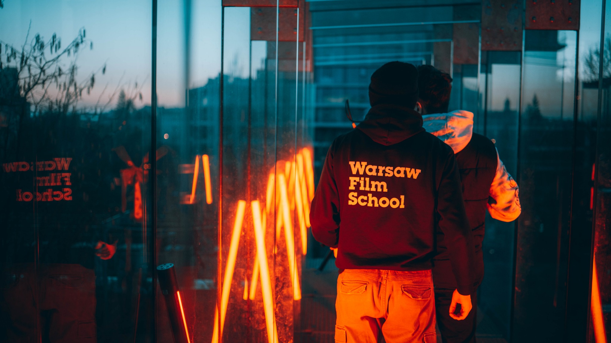 Dwie osoby stojące tyłem, jedna w bluzie z napisem 'Warsaw Film School', oświetlone neonowymi lampami w szklanym labiryncie. Widoczne odbicia budynków i drzew w szklanych ścianach.