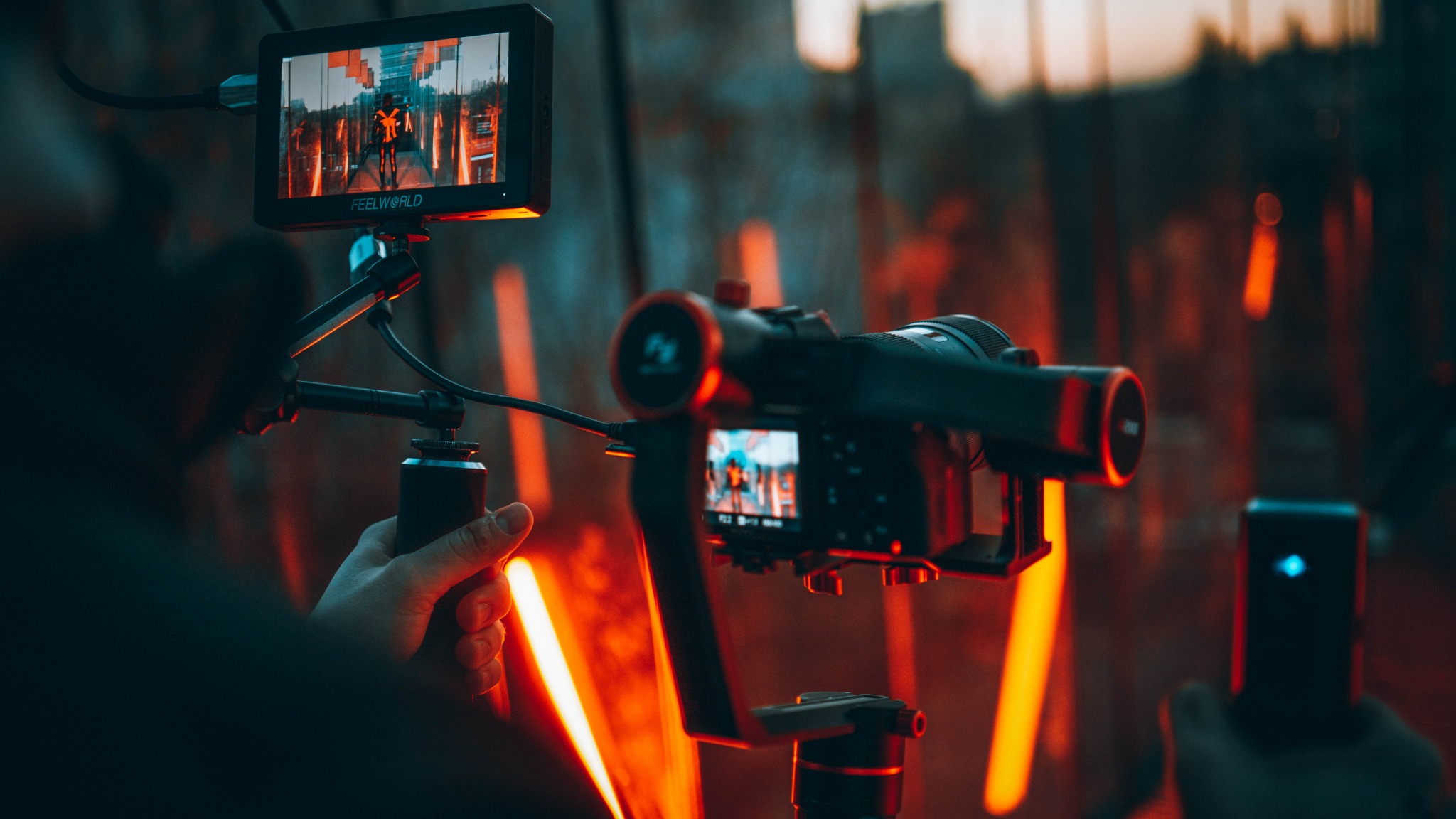 Kamera na stabilizatorze z podglądem nagrywanego ujęcia, dodatkowy monitor Feelworld podłączony kablem, pomarańczowe światła w tle.
