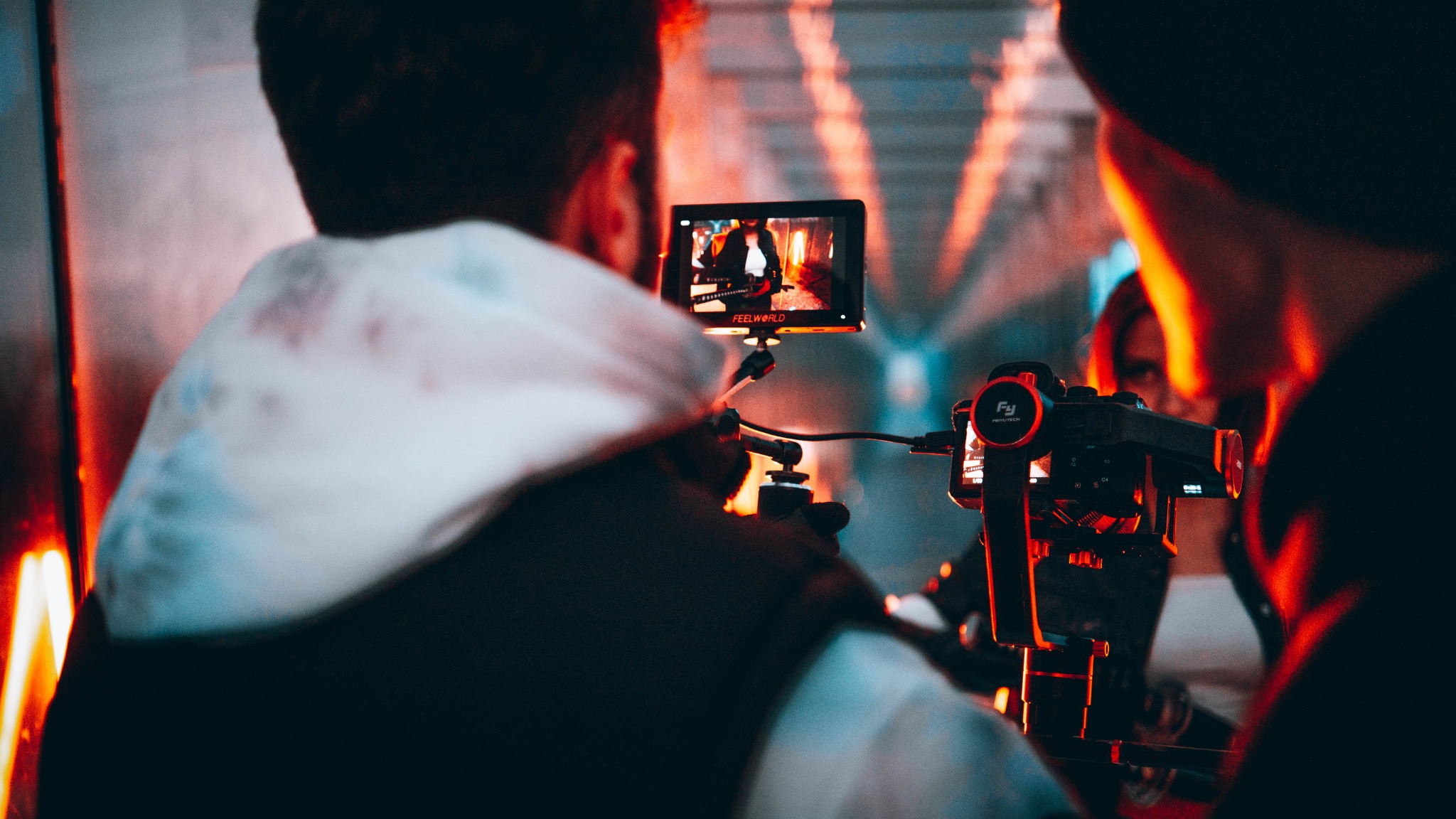 Ekipa filmowa kręci ujęcie w scenerii z neonowym oświetleniem, operator skupiony na monitorze podglądowym stabilizatora obrazu.