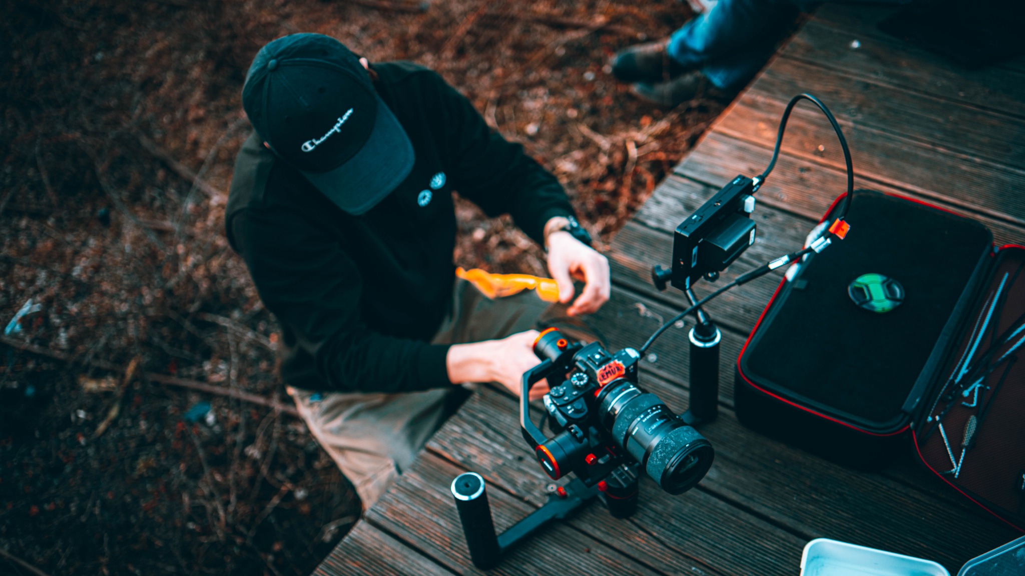 Operator w czarnej bluzie i czapce Champion przygotowuje kamerę z gimbalem na drewnianym pomoście, obok otwarty futerał z akcesoriami i dodatkowy monitor.