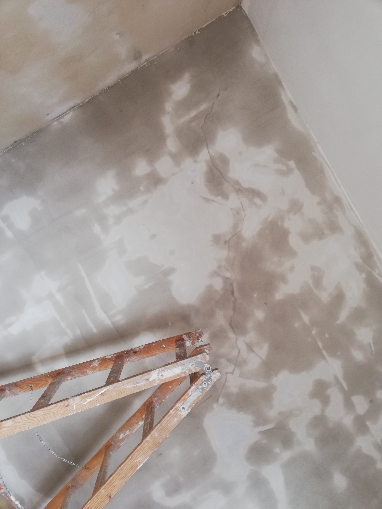 Ściana w trakcie nakładania gładzi, widoczne nierówności i mokre plamy, drabina drewniana oparta o ścianę, sufit pomalowany na biało.