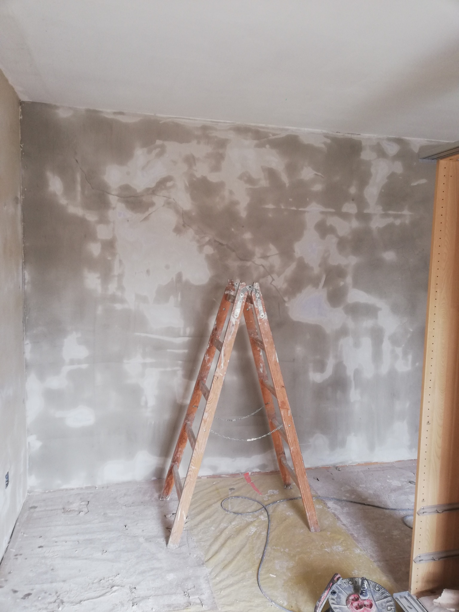 Pokój w trakcie szpachlowania ścian, widoczna drewniana drabina oparta o ścianę z nałożoną warstwą szpachli oraz rozłożona folia ochronna na podłodze.