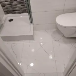 Nowoczesna łazienka po remoncie: kabina prysznicowa z czarno-białymi płytkami na ścianie i biała toaleta na podłodze z płytek imitujących marmur.