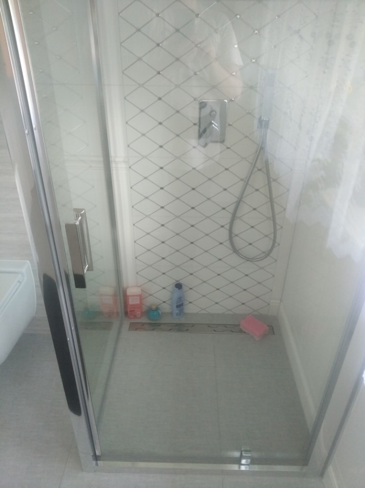 Szklana kabina prysznicowa z chromowanymi okuciami, wyłożona białymi płytkami w romby, widoczny odpływ liniowy, kilka kosmetyków na podłodze i słuchawka prysznicowa.