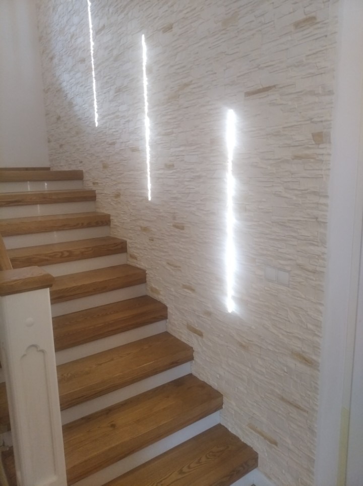 Schody drewniane z białymi podstopnicami i ozdobną balustradą, ściana wyłożona dekoracyjnym kamieniem z wbudowanym oświetleniem liniowym.