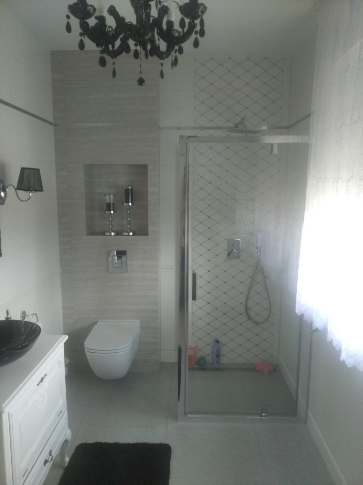 Nowoczesna łazienka z kabiną prysznicową w chromowanej ramie, wiszącą toaletą, umywalką na szafce w stylu retro, czarnym żyrandolem i dekoracyjną wnęką ścienną z ozdobnymi świecznikami.