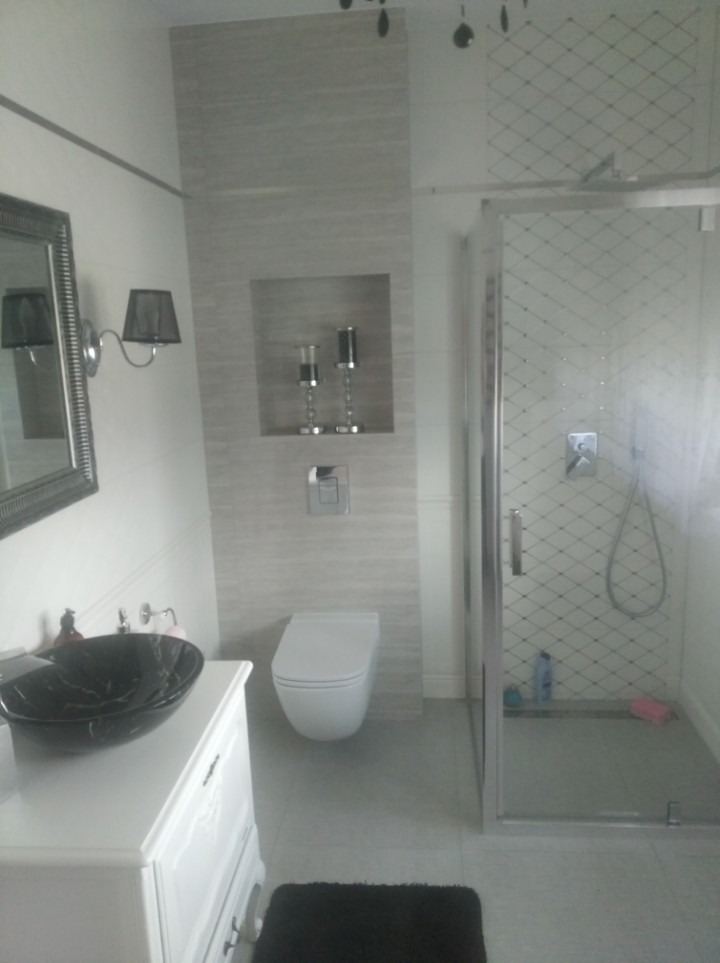 Nowoczesna łazienka z kabiną prysznicową z wzorem rombów, wiszącą toaletą, czarną umywalką na białej szafce, lustrem w ozdobnej ramie i wnęką z dekoracyjnymi świecznikami.