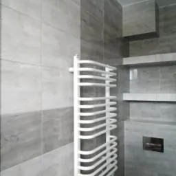 Jasnoszara łazienka z płytkami imitującymi drewno, biały, drabinkowy grzejnik na ścianie, zabudowane półki i ukryta spłuczka.