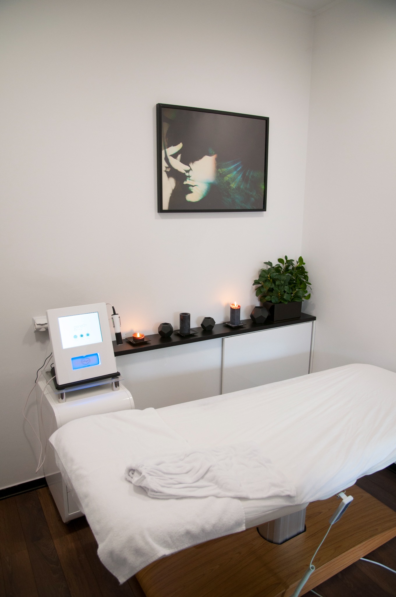 Klimatyczne wnętrze gabinetu zabiegowego z białym łóżkiem, nowoczesnym urządzeniem z ekranem dotykowym, świecami i dekoracyjną grafiką na ścianie.